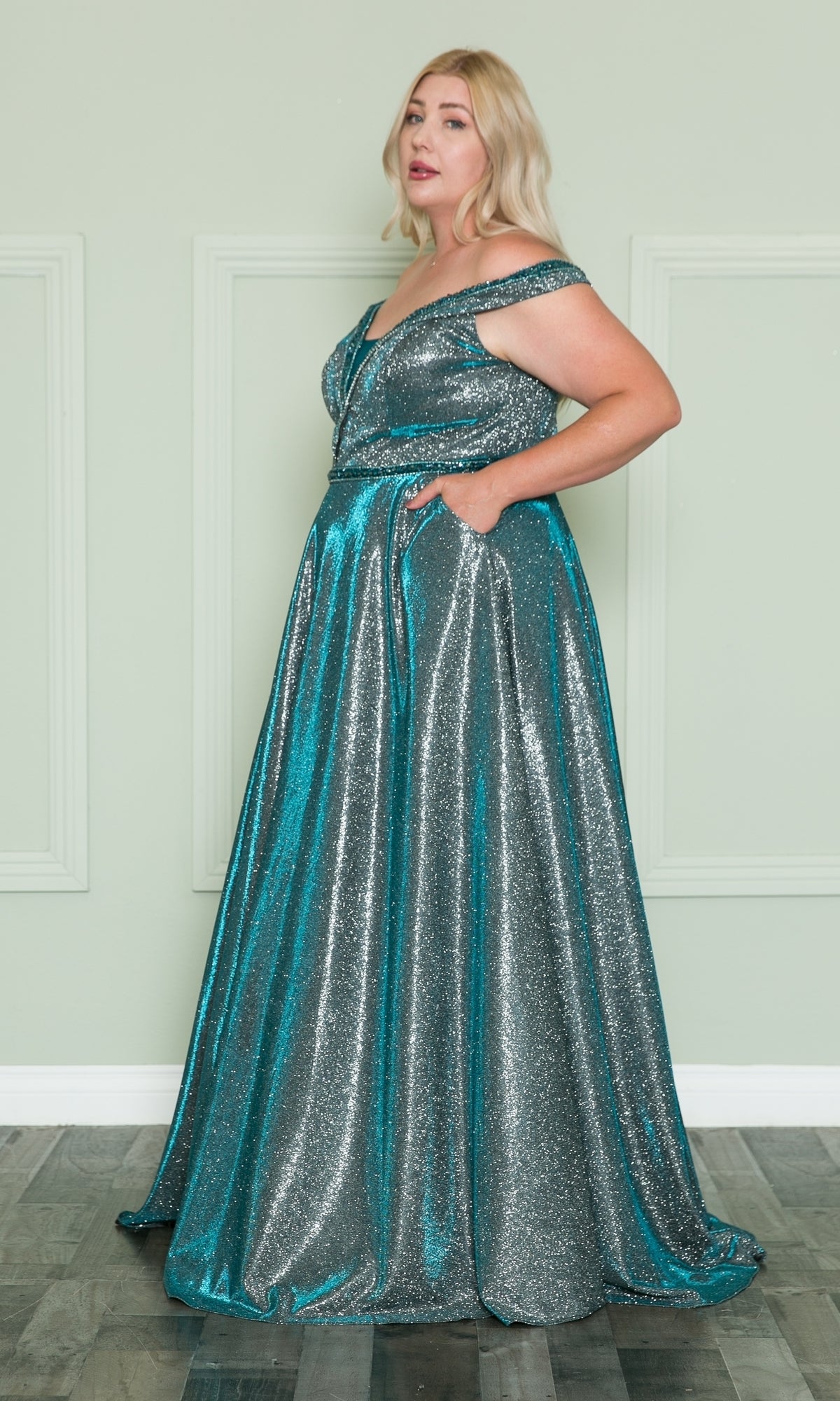 A woman stands before a light green wall, wearing the Plus-Size Long Glitter-Knit Prom Dress W1096—an off-the-shoulder, floor-length teal and silver gown with pockets.